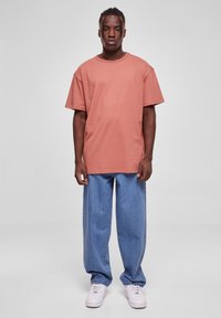 Tricou supradimensionat cu mânecă scurtă, în nuanțe de coral estompat, asociat cu blugi denim lejeri de culoare albastru deschis și adidași albi. Design simplu, fără străluciri.
