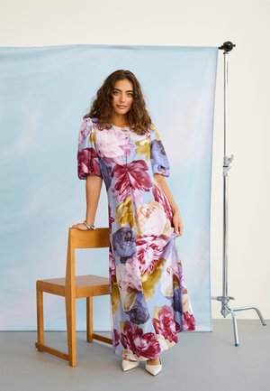 Kobieta o kręconych włosach ubrana w długą, kwiecistą sukienkę z bufiastymi rękawami, stoi obok drewnianego krzesła na jasnoniebieskim tle.