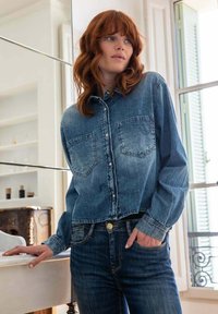 Woman with red hair wearing a denim shirt and jeans, standing indoors by a window with one hand in pocket and the other resting on a table.
