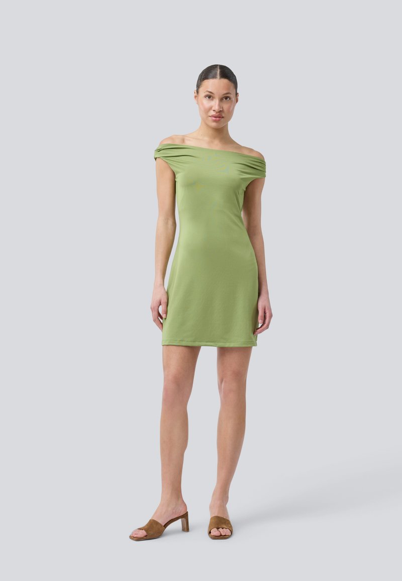 Abito verde off-the-shoulder in tessuto leggero con texture liscia. Presenta una silhouette aderente e arriva sopra la metà della coscia. Sandali con tacco marroni.