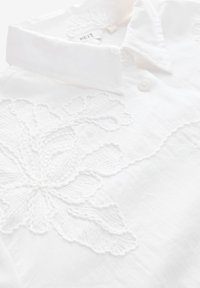 Chemise col blanc avec broderie florale texturée sur le devant, en tissu doux et léger avec fermeture à boutons.