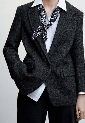 Persona con una chaqueta cruzada de espiga oscura, camisa blanca, pañuelo negro con estampado en el cuello, pantalones negros, mano en el bolsillo, con anillos de oro en la mano derecha.