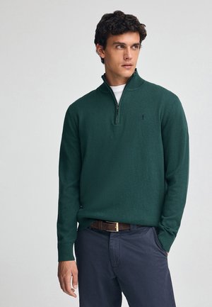 Veste zippée vert avec col en côtelé, manches longues et petit logo. Associée à un pantalon sombre et une ceinture en cuir. Texture lisse.