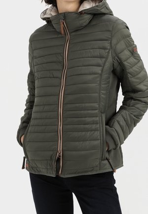 Personne portant une veste matelassée vert olive avec une capuche, une fermeture éclair à l'avant et les mains dans les poches latérales, sur un fond uni.