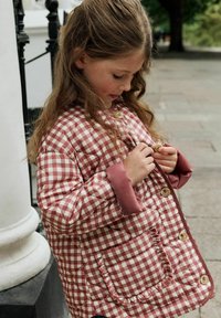 Steppjacke mit rot-weißer karierten Muster, ausgestattet mit einer Rüschentasche und kontrastierenden pinken Manschetten, die mit großen Knöpfen gesichert ist.