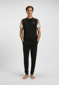 Man staat blote voeten, gekleed in een zwart, bruin en wit kortemouwen shirt en zwarte broek met "BOSS" logo's, voor een effen achtergrond.