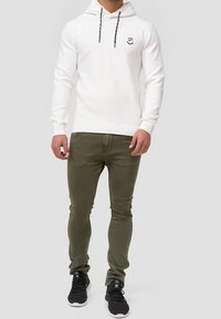 Bílá mikina s kapucí a šňůrkami, žebrovanými manžetami a malým logem. Olivově zelené slim-fit kalhoty. Černé tenisky s texturovanou podrážkou.