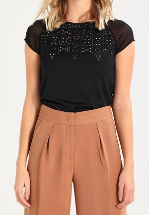 Schwarzes Kurzarmoberteil mit transparentem Schulterdesign und dekorativem Ausschnittmuster. Kombiniert mit hochtaillierten, hellbraunen, gefalteten Hose.