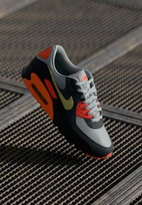 Nike Air Max-sneaker med grå overdel, oransje detaljer, grønn swoosh og teksturert gummisåle med fremtredende luftdemping.