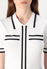 Balta apykaklė marškiniai su juodais akcentais, dviem kišenėmis ir tekstūruotu audiniu. Trumpomis rankovėmis su juodais kraštais ir vertikaliomis detalėmis.