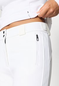 Pantaloni albi cu o texură netedă, având buzunare cu fermoar negre, închidere cu două nasturi și un accent alb la talie.