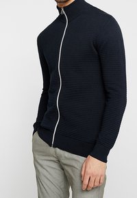 Mörkblå zip-up tröja med ribbad struktur, hög krage och vit dragkedja. Slim fit-design med långa ärmar och kontrasterande muddar.