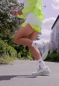 Neon gul atletik shorts og vindjakke parret med hvide sokker og grå-blå løbesko med grønne accenter; grussti og grønt område.