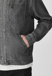 Main dans la poche d'une veste en denim noir délavé avec poches poitrine et latérales, portée sur une chemise blanche et un pantalon noir.