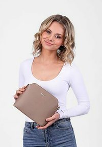 Béžová kožená psaníčka s texturovaným povrchem, se zlatým logem, ostrým tvarem a zipovým uzávěrem. Drží je modelka v ruce.