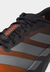 Κοντινή λήψη μαύρου και πορτοκαλί αθλητικού παπουτσιού με μαύρα κορδόνια, τρεις γκρίζες ρίγες στο πλάι και ετικέτα «ADIZERO» στη γλώσσα.