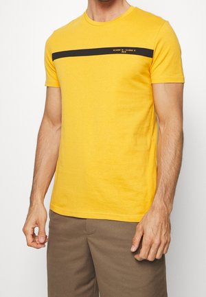 Man wearing a yellow short-sleeve t-shirt with a black horizontal stripe and coordinates printed across the chest, paired with brown pants.