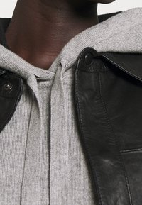 Czarna skórzana kurtka z zatrzaskami, noszona na szarym swetrze z dzianiny z kapturem. Tekstury materiałów kontrastują, a na swetrze znajdują się żebrowane detale.