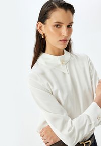 Weiße Bluse mit langen Ärmeln, spitzem Kragen und einem einzelnen Perlenknopf am Hals. Glatte Textur, taillierte Passform und minimalistisches Design.
