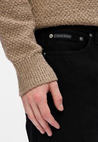 Bežinė megzta palaidinė su gipiūriniais rankogaliais, derinama su juodais džinsais, kuriuose matomas Calvin Klein etiketas ir sidabriniai priedai.