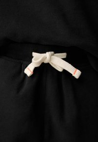 Černá látka od džesíku s bílou šňůrkou svázanou do mašle, na jednom konci s malým oranžovým akcentem. Měkká textura, útulný design.