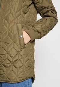 Hand in pocket van olijfgroen gewatteerd jasje met drukknoop en geribbelde manchetten, gecombineerd met blauwe denim jeans op een lichte achtergrond.