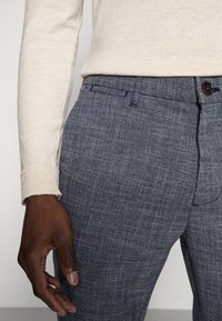 Tmavě modré textured kalhoty s rovný střih, jednoduchým zapínáním na knoflík a jemným vzorkem látky. Světle krémový svetr částečně viditelný.