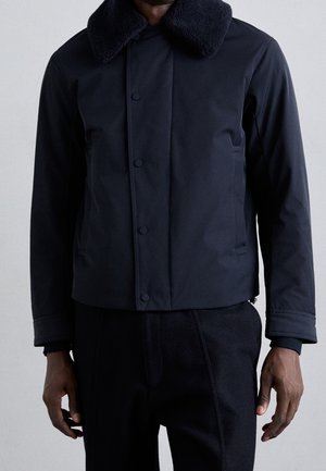 Mann trägt eine dunkelblaue Jacke mit Knopfleiste vorne und einem dicken, strukturierten Kragen, kombiniert mit passenden dunklen Hosen.