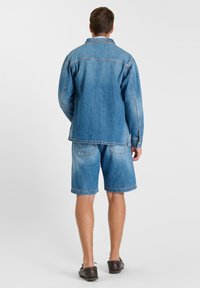 Homme portant une veste en jean bleue et un short en jean assorti, debout de dos sur un fond clair uni.