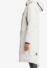 Pitkä valkoinen pufftakki hupulla, jossa on mustat sivuvetoketjulliset taskut, tyylikäs muotoilu ja hillitty rakenne. näkyvät elastiset hihansuut.