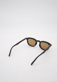 Γυαλιά ηλίου με μαύρο πλαστικό σκελετό και καφέ φακούς, ανοιχτά και τοποθετημένα σε λευκή επιφάνεια, γωνιακά ώστε να φαίνεται η εσωτερική πλευρά του σκελετού.