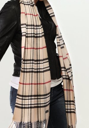 Personne portant une écharpe à carreaux beige avec des franges, une veste en cuir noire, une chemise blanche et un jean bleu sur un fond clair uni.