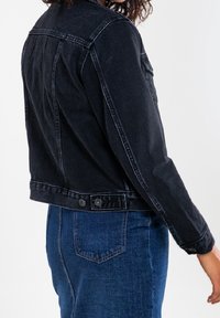 Musta farkkutakki istuvalla leikkauksella, jossa on napillinen etuosan sulkeminen ja sivutaskut. Takana on jäsennelty yoke-muotoilu.