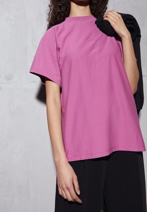 Różowa bawełniana koszulka z krótkimi rękawami i okrągłym dekoltem. Na piersi dyskretne logo. Połączona z czarnym ubraniem.