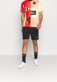Nike fotbollströja i röd, orange och gul med geometriska mönster, i kombination med svarta shorts. Inkluderar logotypdetaljer och vita fotbollsskor.