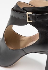 Černá kožená bota na vysokém podpatku s výřezovým designem, sankou kolem kotníku a zlatou přezkou. Hladký povrch a zaoblená špička.