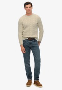 Maglione in cable knit beige chiaro, jeans blu, cintura in pelle marrone e stivaletti stringati marroni. Vestibilità media, polsini a costine e motivo testurizzato.