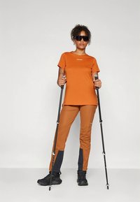 Πορτοκαλί πουκάμισο με κοντά μανίκια και λογότυπο, συνδυασμένο με καφέ παντελόνι με σκούρες γκρι λεπτομέρειες. Μαύρα μπαστούνια πεζοπορίας και παπούτσια ολοκληρώνουν το ντύσιμο.