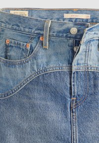 Jupe en denim bleu avec poches avant, rivets en cuivre, passants de ceinture, fermeture par boutons et étiquettes indiquant "Quality Clothing Premium" et taille 27.