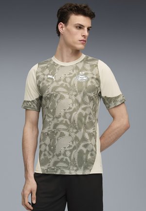 Jongeman met kort donker haar, gekleed in een beige en groen gestreept PSV voetbalshirt en zwarte shorts, staande tegen een grijze achtergrond.