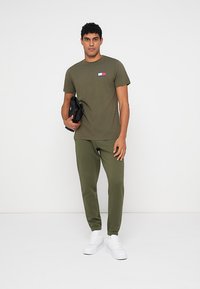 Άνδρας που στέκεται φορώντας ελαιόχρωμο T-shirt και φόρμες, κρατώντας μια μαύρη τσάντα, με λευκά αθλητικά παπούτσια μπροστά σε αφαιρετικό λευκό φόντο.