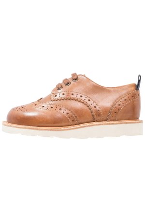 Chaussure brogue en cuir marron avec perforations décoratives, fermeture à lacets à l'avant et semelle blanche, présentée de profil sur fond blanc.