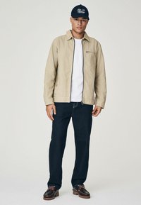 Beige jack met rits en kraag, witte t-shirt, donkerblauwe jeans en donkerbruine schoenen met accenten. Marineblauwe pet met witte tekst.