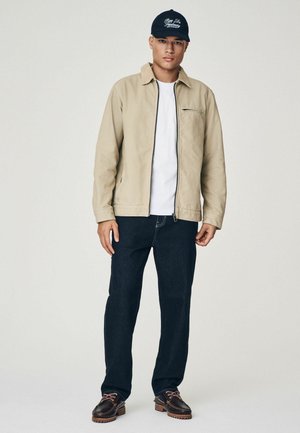 Beige jack met rits en kraag, witte t-shirt, donkerblauwe jeans en donkerbruine schoenen met accenten. Marineblauwe pet met witte tekst.