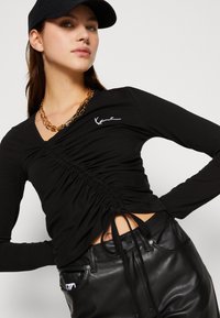 Sort langærmet top med V-hals, der har samlet detaljer og en side snor. Parret med sorte læderbukser og en halskæde i guld.