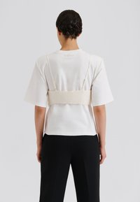 Bēšs bralets ar regulējamām lencēm, valkāts virs baltas īsroku krekla, kombinēts ar melnām pielāgotām biksēm. Gluds audums un minimālistisks dizains.