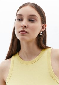 Gul ribbad linne med rund halsringning, kombinerat med silverfärgade hängande örhängen. Slät textur, minimal design, med axelutbyte.