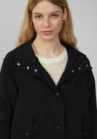Veste noire avec capuche, boutons argentés brillants et poches, superposée à un pull en tricot léger. Texture lisse, design décontracté.