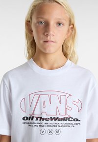 Hvid bomulds t-shirt med rød grafisk tekst "VANS Off The Wall Co." med sorte accenter. Rund hals og korte ærmer.