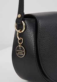 Angolo di una borsa in pelle nera a granuli con anelli dorati e un'etichetta rotonda nera e oro che recita "Coccinelle Authentic Italian Leather".